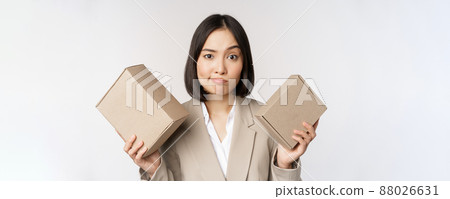 Puzzled asian saleswoman holding boxes, looking confused, standing in business suit over white background Puzzled asian saleswoman holding boxes, looking confused, standing in business suit over white background 88026631