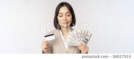Thoughtful businesswoman, korean corporate woman showing credit card and money cash, dollars in hands, standing over white background and thinking 88027919