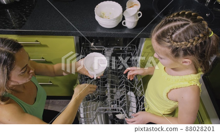 Mother teaching daughter smart girl learning to use dishwasher. Young mistress children loading putting dirty dishes in automatic dishwasher. Mother teaching daughter smart girl learning to use dishwasher. Young mistress children loading putting dirty dishes in automatic dishwasher. 88028201