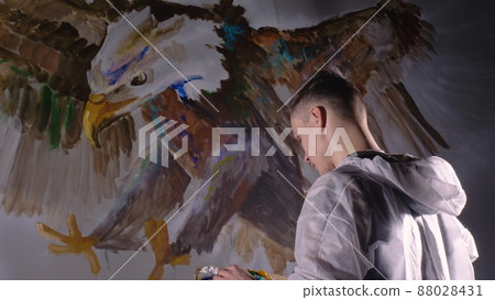 Artist designer draws an eagle on wall. Craftsman decorator paints picture with acrylic oil color looking at sketch in phone. Dark magic cinematic. Artist designer draws an eagle on wall. Craftsman decorator paints picture with acrylic oil color looking at sketch in phone. Dark magic cinematic. 88028431
