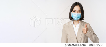 Coronavirus and workplace concept. Portrait of businesswoman in medical face mask showing thumbs up, wearing business suit, standing over white background 88029533