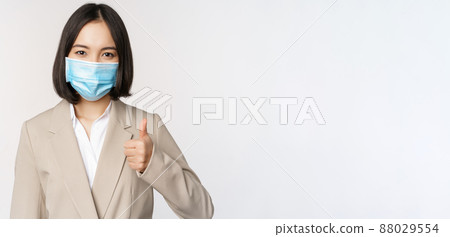 Coronavirus and workplace concept. Portrait of businesswoman in medical face mask showing thumbs up, wearing business suit, standing over white background Coronavirus and workplace concept. Portrait of businesswoman in medical face mask showing thumbs up, wearing business suit, standing over white background 88029554