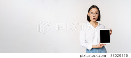 Enthusiastic asian woman, office worker in glasses showing digital tablet screen, demonstrating info on gadget display, standing over white background Enthusiastic asian woman, office worker in glasses showing digital tablet screen, demonstrating info on gadget display, standing over white background 88029556