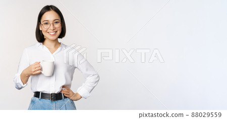 Happy young energetic asian woman smiling, drinking, holding cup mug of coffee, standing confident against white background Happy young energetic asian woman smiling, drinking, holding cup mug of coffee, standing confident against white background 88029559