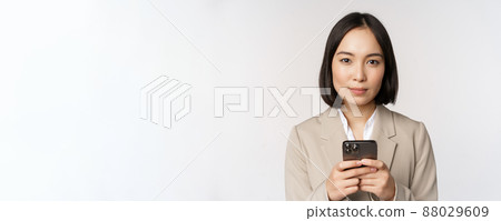 Image of asian businesswoman in suit, holding mobile phone, using smartphone app, smiling at camera, white background 88029609