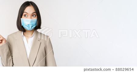 Enthusiastic businesswoman pointing fingers left, wearing medical face mask from covid-19 pandemic, standing over white background Enthusiastic businesswoman pointing fingers left, wearing medical face mask from covid-19 pandemic, standing over white background 88029654