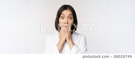 Close up face of asian woman gasping, looking shocked and speechless, holding hands near mouth and staring at camera impressed, white background 88029749