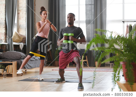 Sporty man and woman doing strengthening exercises in loft living room using resistance band and dumbbells Sporty man and woman doing strengthening exercises in loft living room using resistance band and dumbbells 88033030