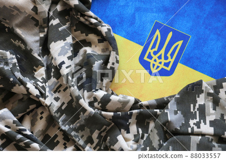Ukrainian flag and coat of arms with fabric with texture of pixeled camouflage. Cloth with camo pattern in grey, brown and green pixel shapes with Ukrainian trident sign Ukrainian flag and coat of arms with fabric with texture of pixeled camouflage. Cloth with camo pattern in grey, brown and green pixel shapes with Ukrainian trident sign 88033557