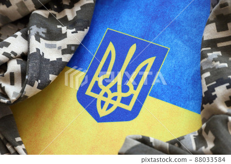 Ukrainian flag and coat of arms with fabric with texture of pixeled camouflage. Cloth with camo pattern in grey, brown and green pixel shapes with Ukrainian trident sign 88033584