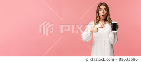 Have you seen it, awesome app. Impressed and excited pretty blond girl in white dress recommend download application, visit online store, pointing at mobile phone and stare camera thrilled 88033680