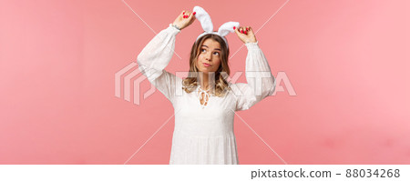 Holidays, spring and party concept. Portrait of silly cute blond girl in rabbit ears, looking up daydreaming, smiling lovely wear white dress, standing over pink background carefree 88034268