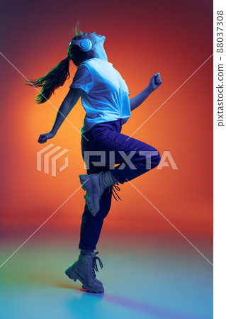 Young happy girl emotively listening to music in headphones and dancing isolated over gradient red yellow background in neon ligh Young happy girl emotively listening to music in headphones and dancing isolated over gradient red yellow background in neon ligh 88037308