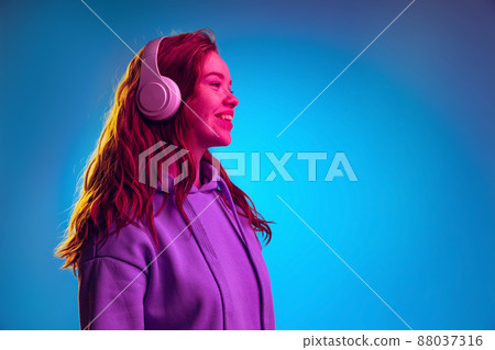 Side view portrait of young girl in white headphones posing isolated over blue studio background in pink neon light Side view portrait of young girl in white headphones posing isolated over blue studio background in pink neon light 88037316