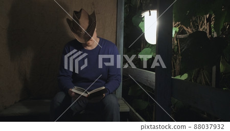 Cowboy man is reading a paper book sitting on the porch of a village house. The concept of reading paper books, learning and hobbies. Dark night. Old, retro decor, burlap background Cowboy man is reading a paper book sitting on the porch of a village house. The concept of reading paper books, learning and hobbies. Dark night. Old, retro decor, burlap background 88037932