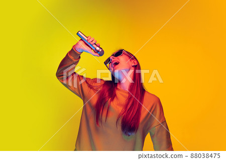 Portrait of young girl singing in microphone, posing isolated over yellow studio background in neon light Portrait of young girl singing in microphone, posing isolated over yellow studio background in neon light 88038475