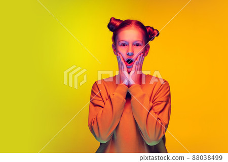 Portrait of red haired teen girl posing with shocked emotions isolated over yellow studio background in neon light 88038499