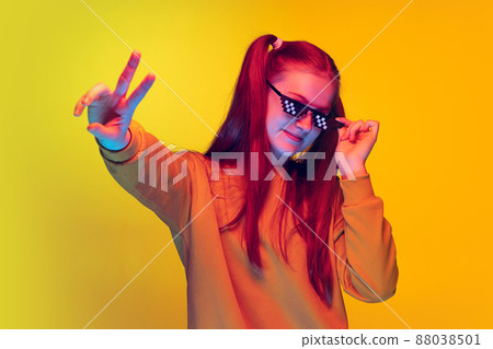 Portrait of teen girl in pixel sunglasses showing peace sign with fingers, posing isolated over yellow studio background Portrait of teen girl in pixel sunglasses showing peace sign with fingers, posing isolated over yellow studio background 88038501