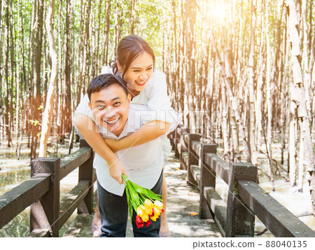An Asian woman in a white dress holding a bouquet of flowers happily rides on the back of an Asian man in the mangrove forest. To show love and happiness after being proposed with cool shade 88040135