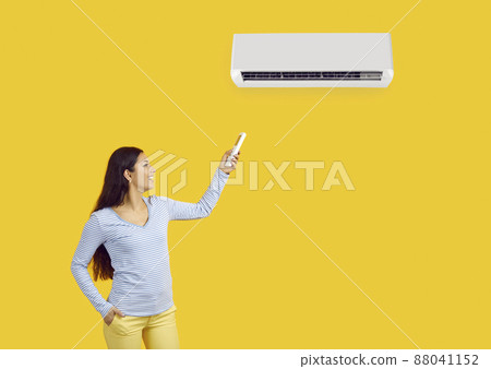 Woman with remote control adjusts comfortable temperature on air conditioner on yellow background. Young happy woman uses modern air conditioning for her comfort. Banner. Copy space. Advertising. 88041152