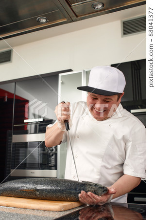 The big salmon is in the hands of the asian chef cook. He is using a knife to slice salmon fillet. 88041377