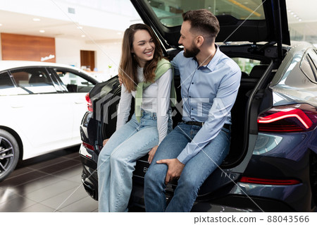 happily young couple sitting in car trunk dangling legs in car dealership 88043566