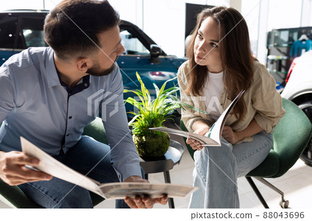 a young couple in a car dealership studying booklets with specifications and prices for new cars a young couple in a car dealership studying booklets with specifications and prices for new cars 88043696