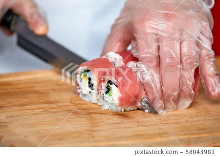 Chef cuts sushi rolls with red salmon fish with a knife on a wooden board. Chef in transparent gloves works. 88043981