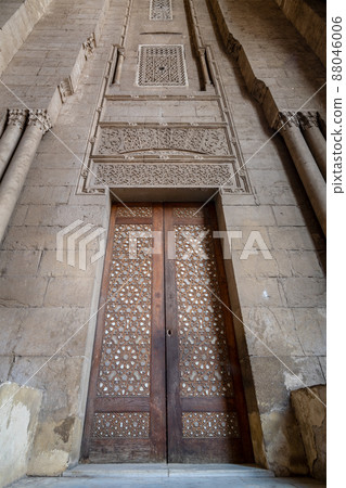 External old decorated bricks stone wall with arabesque decorated wooden door framed by stone ornate cylindrical columns 88046006