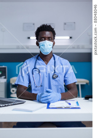 Healthcare facilty nurse sitting at desk while wearing facemask. Clinic nurse sitting at office table in clinic workspace while wearing surgical gloves and virus protection mask. Healthcare facilty nurse sitting at desk while wearing facemask. Clinic nurse sitting at office table in clinic workspace while wearing surgical gloves and virus protection mask. 88048046