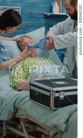 Doctor and nurse using oxygen tube for resuscitation with heart rate monitor in nursing home. Medical team helping retired woman with IV drip bag in hospital bed. Specialists giving support Doctor and nurse using oxygen tube for resuscitation with heart rate monitor in nursing home. Medical team helping retired woman with IV drip bag in hospital bed. Specialists giving support 88048182