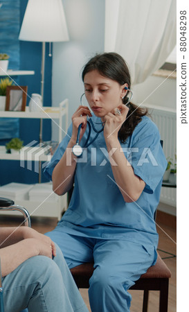 Retired woman in wheelchair receiving consultation from nurse. Medical assistant using stethoscope for healthcare checkup in nursing home. Patient with disability getting assistance. 88048298