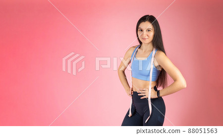 The trainer holds in his hands a measuring tape to measure the body. Attractive brunette woman in fashionable sportswear on pink background. Healthy lifestyle concept The trainer holds in his hands a measuring tape to measure the body. Attractive brunette woman in fashionable sportswear on pink background. Healthy lifestyle concept 88051563
