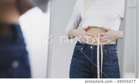 A young woman stands in front of a mirror and measures her waist circumference with a tape measure A young woman stands in front of a mirror and measures her waist circumference with a tape measure 88056877