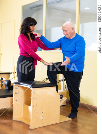 Hispanic woman pilates instructor helping senior man exercising on combo chair Hispanic woman pilates instructor helping senior man exercising on combo chair 88061423