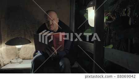 An angry elderly man is sitting on porch at night, he is reading a book. Concept of a scary fairy tale, horror, nightmares and black humor. Old, retro decor, burlap background 88069042
