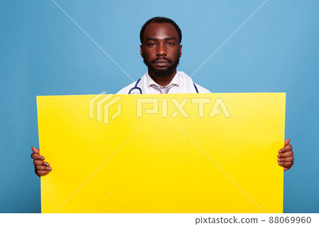 Confident medic with stethoscope holding large yellow empty banner for health care medical concept over blue background. Doctor in white lab coat presenting pannel with advertising copy space. Confident medic with stethoscope holding large yellow empty banner for health care medical concept over blue background. Doctor in white lab coat presenting pannel with advertising copy space. 88069960