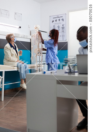 African american specialist nurse with face mask to prevent infection with covid19 showing bone structure skeleton explaining spinal pain to senior old woman. Medical examination in hospital office African american specialist nurse with face mask to prevent infection with covid19 showing bone structure skeleton explaining spinal pain to senior old woman. Medical examination in hospital office 88070016