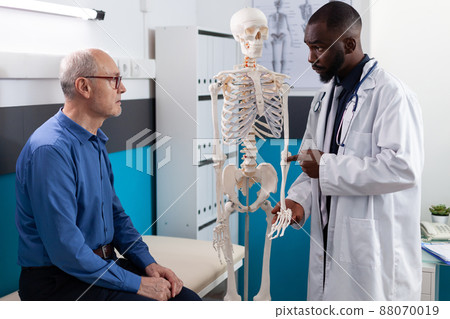 Physiotherapist doctor showing anatomical human skeleton explaining back pain to old man patient discussing sickness symptoms during medical physiotherapy in hospital office. Osteoporosis treatment Physiotherapist doctor showing anatomical human skeleton explaining back pain to old man patient discussing sickness symptoms during medical physiotherapy in hospital office. Osteoporosis treatment 88070019