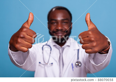 Closeup of hands giving thumbs up of cheerful african american doctor in medical uniform with stethoscope. Cropped detail of optimsitic medic showing hand gesture for positive attitude. Closeup of hands giving thumbs up of cheerful african american doctor in medical uniform with stethoscope. Cropped detail of optimsitic medic showing hand gesture for positive attitude. 88070038