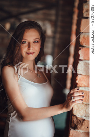 beautiful pregnant woman in white dress by the window with brickwork on walls.  88070888