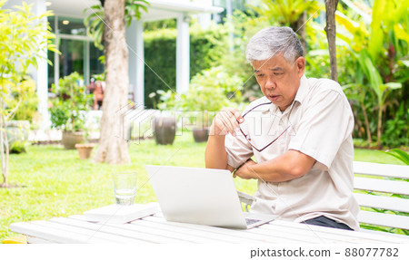 Portrait of old elderly Asian man using a computer laptop in the backyard for learning new skill after retired. Concept of no Ageism and not be late for learning. 88077782
