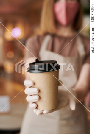 Close-up selective focus on foreground shot of unrecognizable waitperson wearing mask and gloves giving cup of coffee to customer Close-up selective focus on foreground shot of unrecognizable waitperson wearing mask and gloves giving cup of coffee to customer 88079396