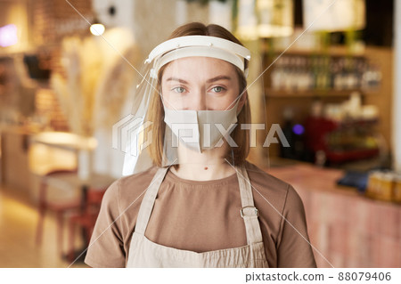 Medium close-up portrait shot of young female cafe worker wearing two kinds of protective mask on face looking at camera Medium close-up portrait shot of young female cafe worker wearing two kinds of protective mask on face looking at camera 88079406