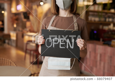 Horizontal medium section shot of unrecognizable woman working in cafe standing indoors holding sign with word Open and protective mask Horizontal medium section shot of unrecognizable woman working in cafe standing indoors holding sign with word Open and protective mask 88079407