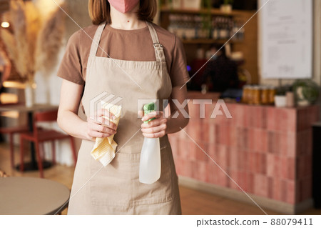 Horizontal medium section shot of unrecognizable young female waitperson wearing mask holding sanitizing spray and wiper Horizontal medium section shot of unrecognizable young female waitperson wearing mask holding sanitizing spray and wiper 88079411