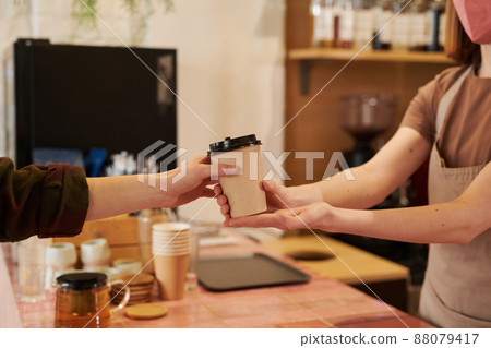 Medium section side view shot of unrecognizable female barista wearing mask standing at counter giving cup of coffee to customer Medium section side view shot of unrecognizable female barista wearing mask standing at counter giving cup of coffee to customer 88079417