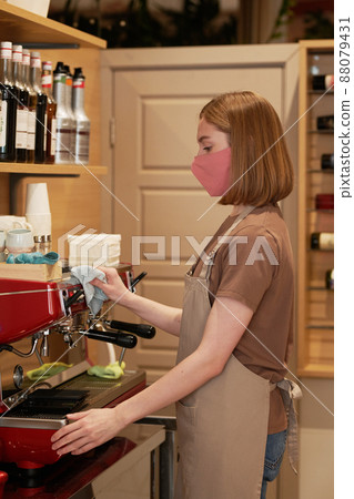 Vertical medium shot of young female cafe wearing protective mask on face and apron cleaning coffee machine 88079431