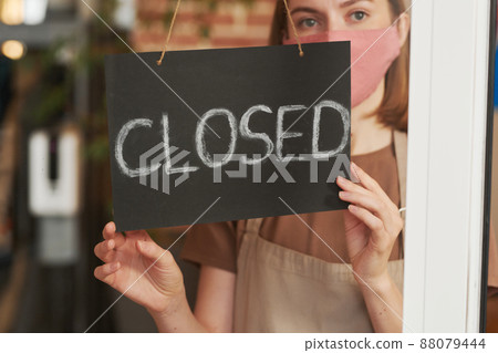 Horizontal medium close-up portrait of young Caucasian woman wearing protective mask on face closing cafe for cleaning it Horizontal medium close-up portrait of young Caucasian woman wearing protective mask on face closing cafe for cleaning it 88079444