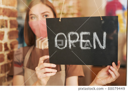Horizontal through glass door portrait of young waitress wearing protective mask on face starting workday with opening cafe Horizontal through glass door portrait of young waitress wearing protective mask on face starting workday with opening cafe 88079449